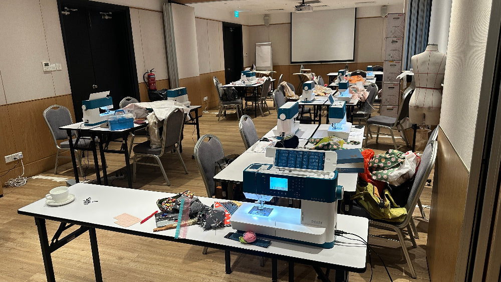 Room with tables and chairs, including 11 sets of PFAFF® ambition™ 620 sewing machine on tables.